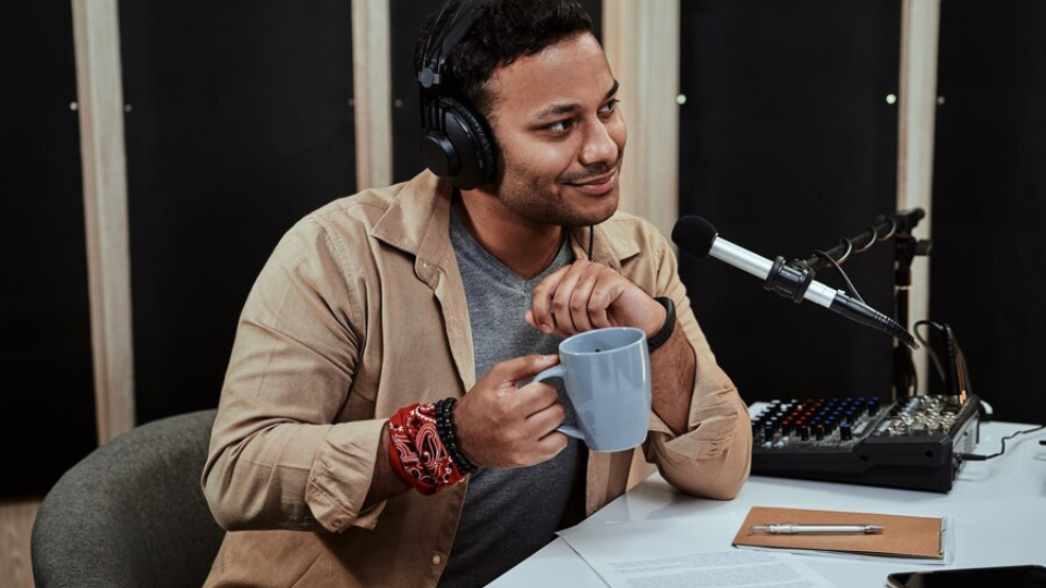 A radio host speaking into a microphone while holding a coffee mug in a studio setting.